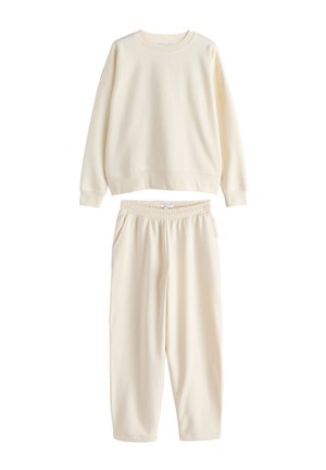 Donna con lunghi capelli scuri che indossa una felpa color crema, pantaloni abbinati, camicia rosa a righe sotto e scarpe slip-on bianche, appoggiata a un muro.