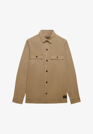 Herrebeige skjorte med lange ærmer og knapper, krave, to brystlommer med knapklapper foran og en lille sort mærkat nær den nederste kant.