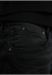 Vaqueros de mezclilla negra con una vista en primer plano del bolsillo, con herrajes plateados, una textura sutil y un detalle en la costura que se asemeja a una "A".