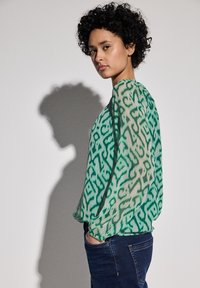 Grün gemusterte Bluse mit transparenter Textur, die ein abstraktes Design aufweist, lange Ärmel und einen runden Halsausschnitt hat, kombiniert mit dunklen Jeans.