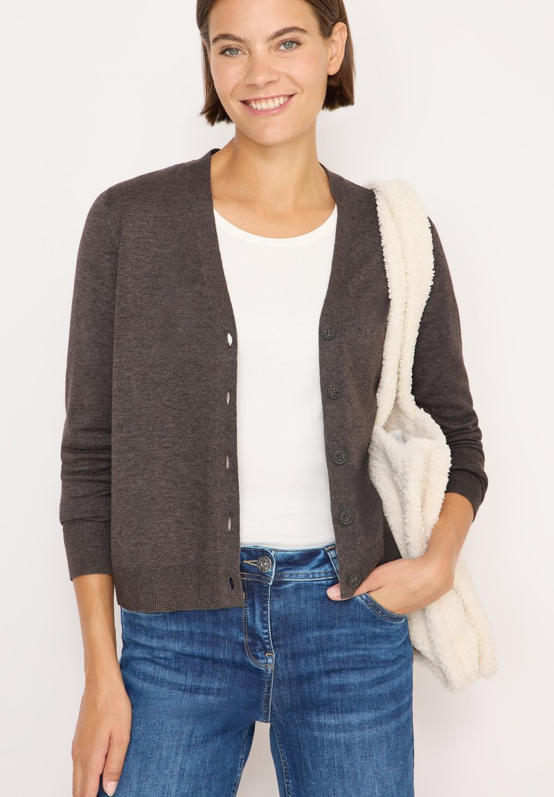 Cardigan marrone lavorato a maglia con scollo a V, cinque bottoni e polsini a coste. Indossato sopra una maglietta bianca, abbinato a jeans blu e una borsa pelosa.