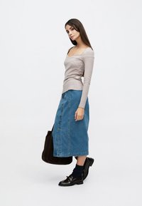 Νεαρή γυναίκα που φοράει μπεζ μπλούζα με μακριά μανίκια, midi φούστα από μπλε τζιν με κουμπιά, μαύρα παπούτσια και κρατάει μεγάλη σκούρα καφέ τσάντα, στέκεται πλευρικά.