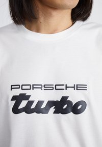 Balta kokvilnas T-kreklu ar melnu tekstu: "PORSCH" treknā fontā virs "turbo" noapaļotā, stilizētā dizainā.
