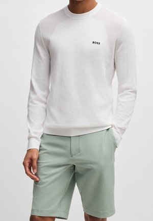 Mand iført en hvid BOSS sweater med lange ærmer og salviegrønne knælange shorts, stående med den ene hånd i lommen.