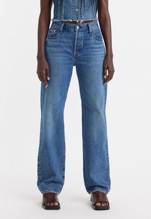 Personne portant un jean bleu taille haute à jambes droites, un gilet en denim court et des sandales noires à talons blocs ouverts à l’orteil sur un fond uni.
