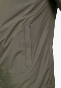 Veste vert olive présentant une texture lisse et élégante, avec une poche dissimulée sur le côté gauche et des détails de couture minimalistes.