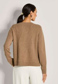 Frau mit dunklem Haar in einem Pferdeschwanz, die einen strukturierten braunen Pullover und weiße Hosen trägt, von hinten vor einem schlichten hellen Hintergrund gezeigt.