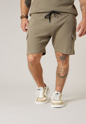 Homme portant un short côtelé beige avec cordon de serrage, des baskets beige et blanches, avec des tatouages visibles sur les jambes et les avant-bras.