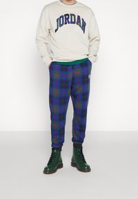 Felpa color crema con il testo "JORDAN" in tonalità scure, abbinata a pantaloni della tuta a quadri blu con righe rosse e verdi. Rifiniture nere sui polsini, stivali verdi.