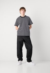 Tricou cu dungi negre și albe, cu mâneci scurte, asortat cu pantaloni din denim închis și teniși albi, creând un aspect casual.