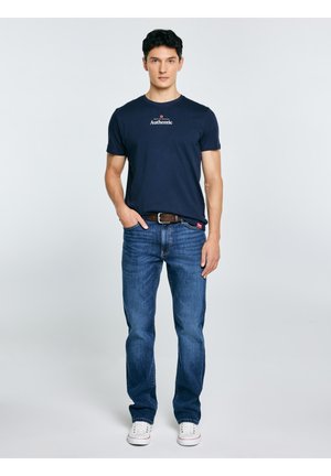 Młody mężczyzna stojący z jedną ręką w kieszeni, ubrany w granatową koszulkę z napisem „Authentic”, niebieskie jeansy, brązowy pasek i białe trampki na jednolitym tle.