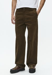 Bruine corduroy broek met een rechte snit, voorzien van een sluiting met knoop aan de voorkant en zakken. Gecombineerd met bruine veterschoenen.
