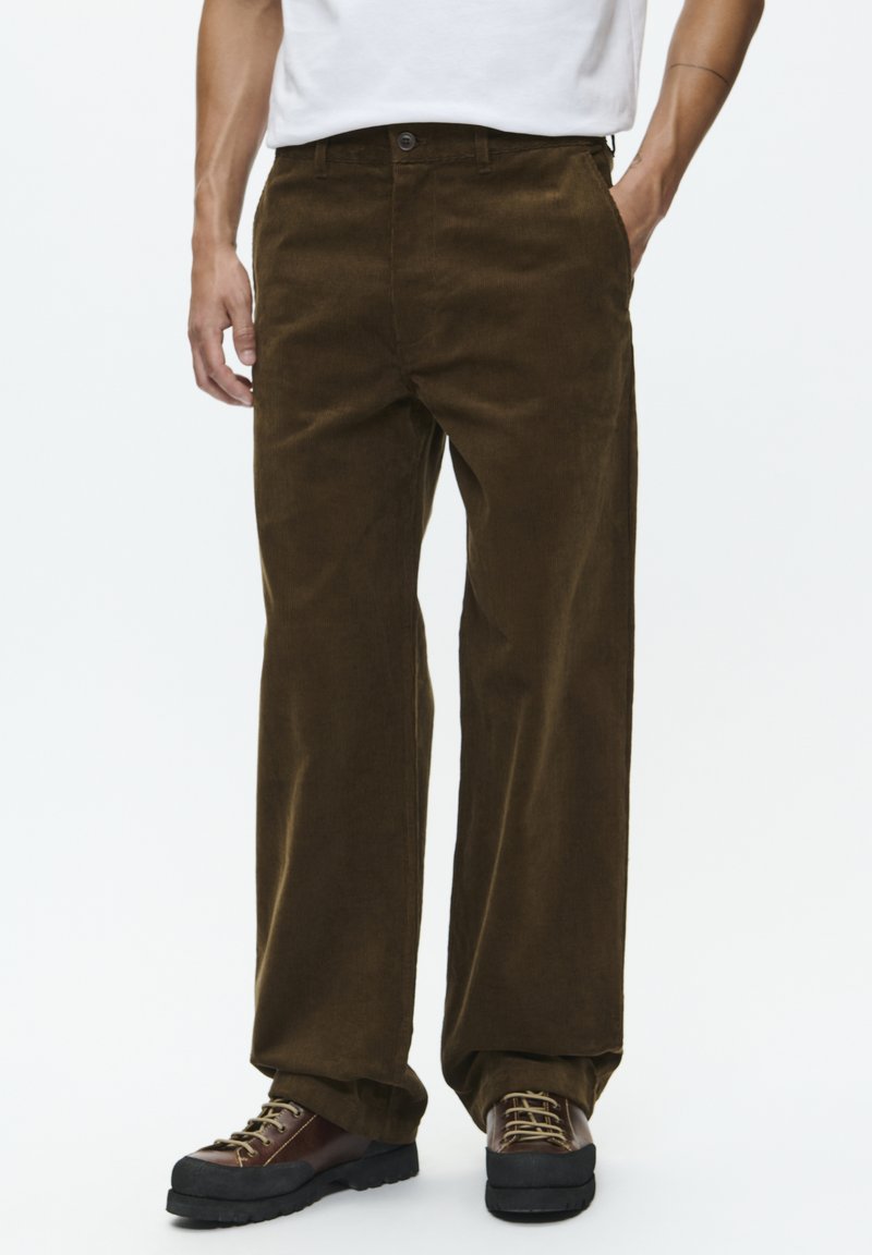 Bruine corduroy broek met een rechte snit, voorzien van een sluiting met knoop aan de voorkant en zakken. Gecombineerd met bruine veterschoenen.