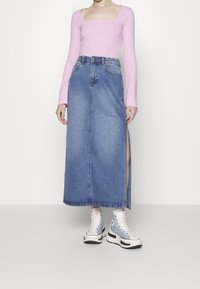 Langer Jeansrock mit seitlichem Schlitz, kombiniert mit einem figurbetonten, rosa Langarmoberteil und hellblauen High-Top-Sneakers. Glatte Texturen, lässiger Stil.
