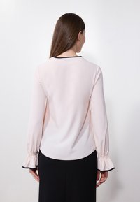 Blusa rosa chiaro con maniche lunghe, polsini arricciati e bordo nero attorno al collo. Tessuto morbido con un taglio dritto e design sul retro.