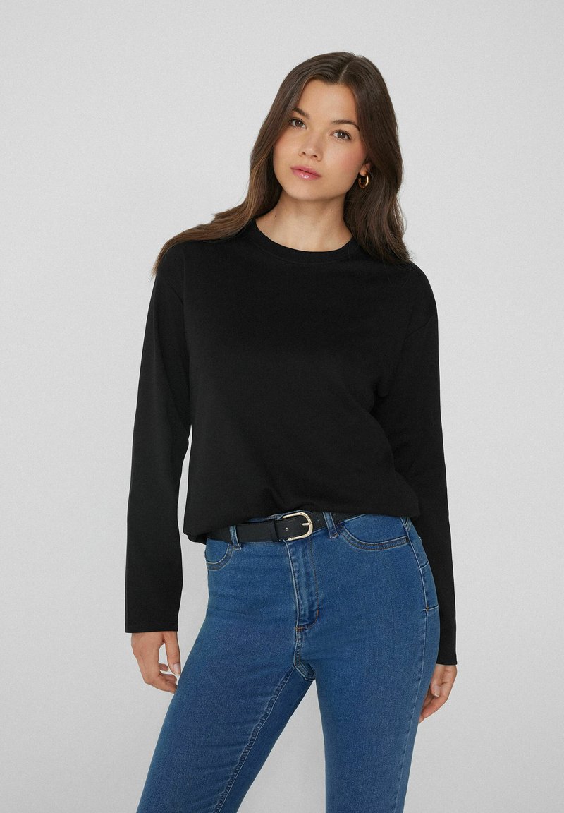 Schwarzes langärmliges T-Shirt mit Rundhalsausschnitt, aus glattem Stoff. Kombiniert mit hochgeschnittenen blauen Jeans und einem schwarzen Gürtel.
