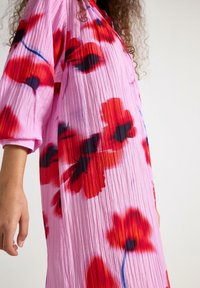 Lyserød plisseret kjole med store røde og sorte blomsterprint, trekvartlange ærmer og en løs pasform.