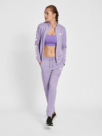 Lavendel tracksuit med en zip-up jakke med hvide striber på ærmerne, parret med en matchende cropped top og joggerbukser. Sorte sneakers.