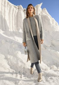 Frau in einem langen grauen Mantel und einem Sweater, dunklen Hosen und beigen Stiefeln, die eine metallische Handtasche hält. Sie steht auf schneebedecktem Boden unter einem klaren blauen Himmel.