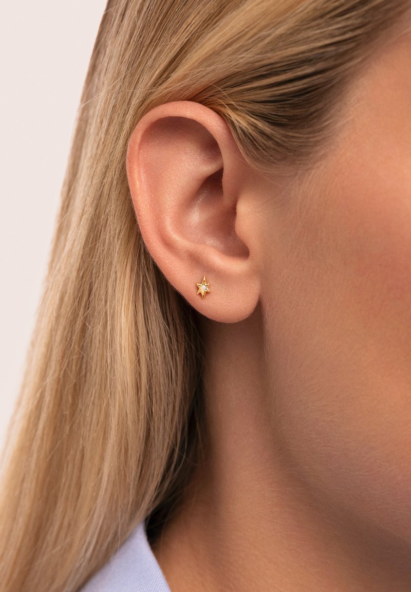 Pendiente de aro en forma de estrella dorada con un pequeño centro de gema, contrastando con un primer plano de un tono de piel clara y cabello rubio liso.