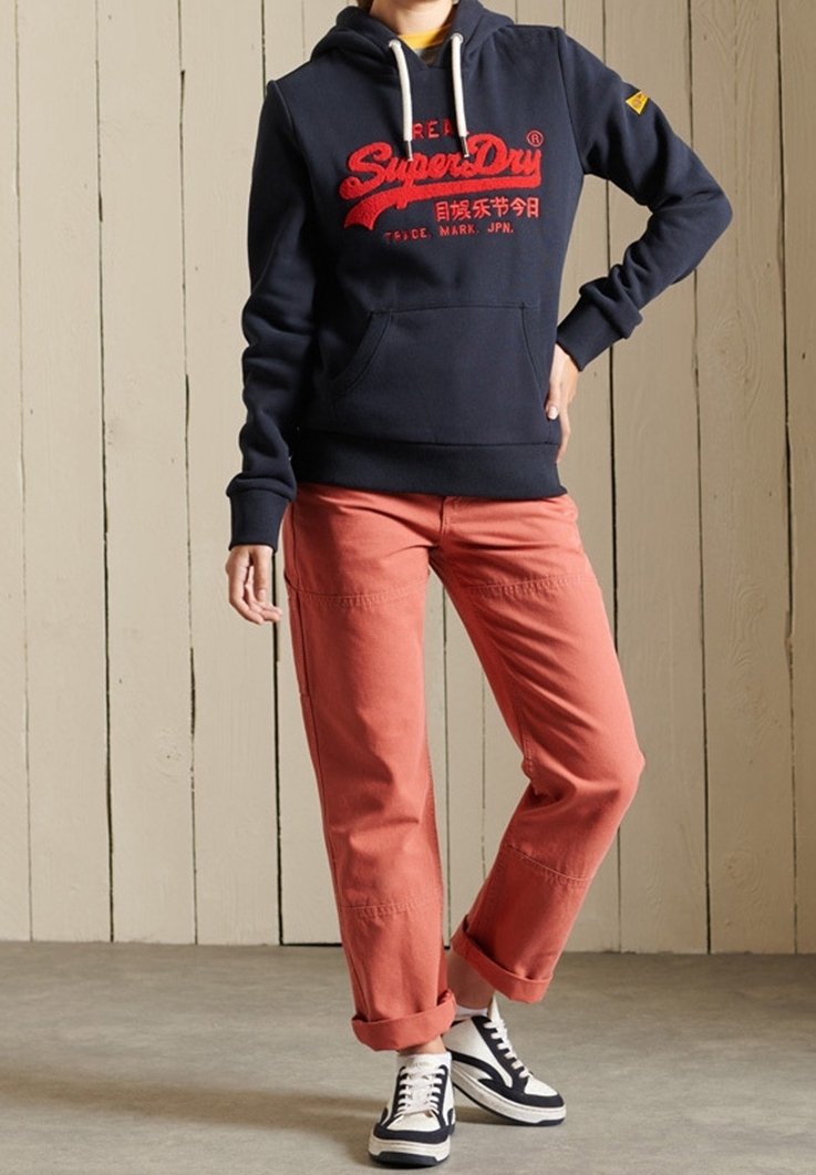 Personne portant un sweat à capuche bleu marine avec du texte rouge, un pantalon rose retroussé et des baskets noires et blanches, debout devant un mur en bois clair.