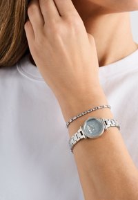 Lacătul unei femei poartă un ceas cu curea din link-uri de argint, cu un cadran texturat și o brățară delicată din argint cu pietre rectangulare.