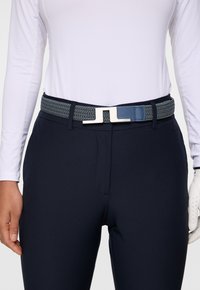 Person som bär marinblå byxor och ett flätat blått bälte med silverspänne, klädd i en vit långärmad skjorta och vit golfhandske på höger hand.