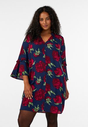 Donna con capelli ricci che indossa un vestito blu largo con grandi motivi floreali rossi e verdi e maniche a campana tre quarti.