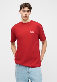 Mladić s kratkom tamnom kosom u crvenoj majici s natpisom "the new originals" i svijetlo plavim trapericama, stoji ispred bijele pozadine.