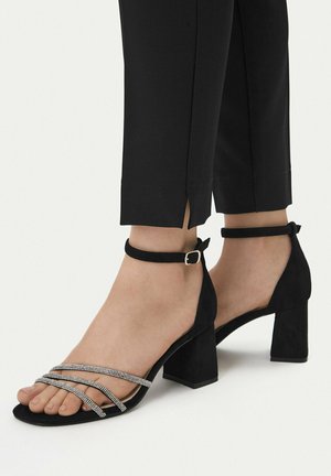 Piedi con sandali neri dal tacco a blocco, cinturini con strass e fibbie alla caviglia, abbinati a pantaloni neri alla caviglia con spacchi laterali.