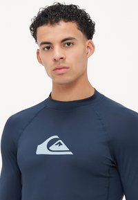 Tengeri kék, hosszú ujjú rash guard, testhezálló kialakítással, rugalmas anyagból készült. A mellkasán világoskék logó található, valamint lapos varrásokkal rendelkezik.
