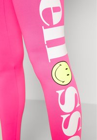 Fukcyjna legginsy w kolorze fuksji z dużym białym napisem "cześć" oraz grafiką żółtej uśmiechniętej buźki, wykonane z elastycznego, gładkiego materiału.