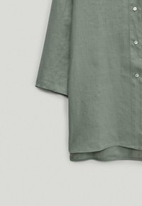 Chemise en lin vert clair avec ouverture à boutons, manches courtes et ourlet arrondi. Présente une texture subtile et des boutons blancs simples.