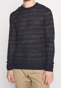Pull en maille marine avec des motifs de texture horizontaux en fils clairs et foncés, doté d'un col rond et de poignets et d'un ourlet en côtes.