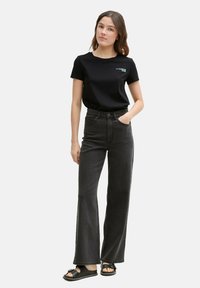 Tricou negru cu logo, asortat cu blugi gri închis cu talie înaltă și sandale negre. Modelul stă în picioare, cu mâinile în buzunare, pe un fundal simplu.