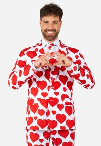 Biely oblek zdobený veľkými červenými srdcovými vzormi, s dvojitým zapínaním a zodpovedajúcimi nohavicami. Model vytvára tvar srdca rukami.