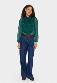 Blouse verte transparente avec un devant à volants, associée à un jean en denim bleu à taille haute. Une ceinture marron et des chaussures marron complètent la tenue.