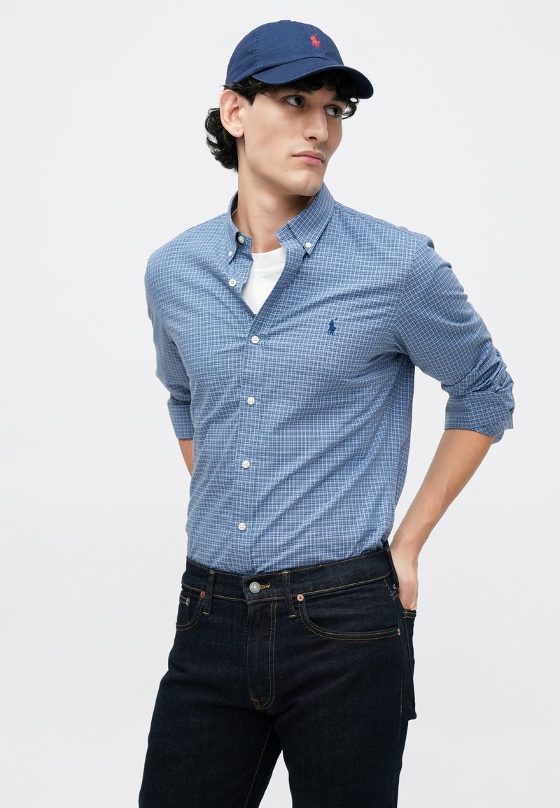 Blåt ternet skjorte med knap og krave samt en hvid undertrøje, parret med mørke denim jeans og en marineblå kasket med et logo.