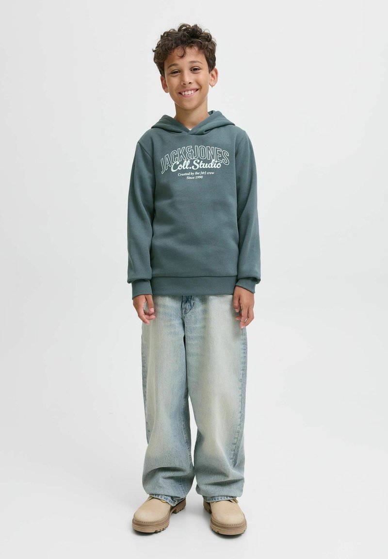 Ragazzino sorridente con i capelli ricci, indossando un hoodie verde Jack & Jones, jeans larghi blu chiaro e stivali beige, in piedi su uno sfondo bianco.