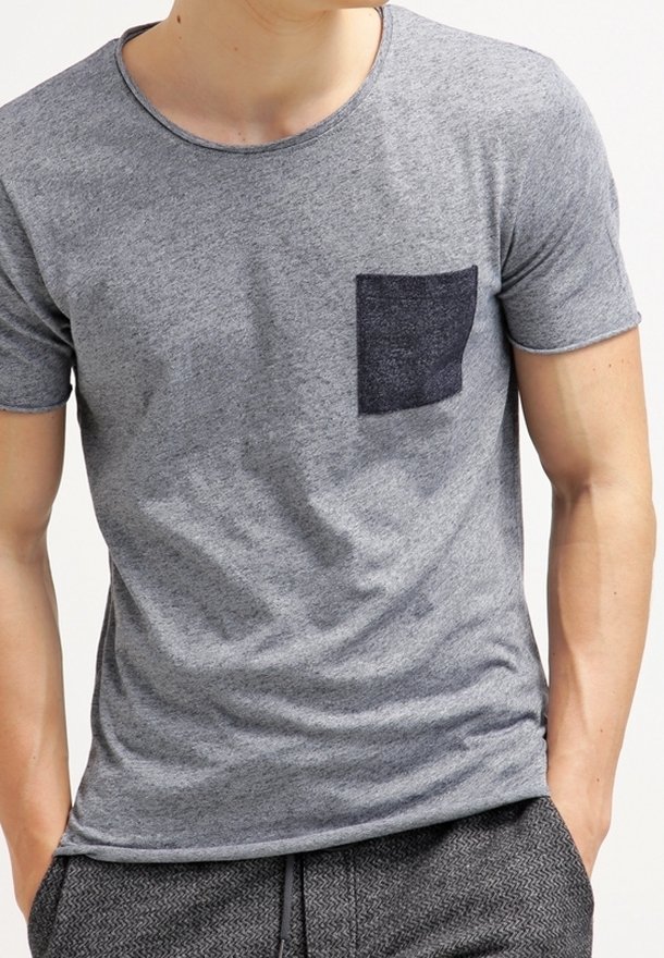 Mężczyzna ubrany w szary t-shirt z krótkim rękawem, z ciemnoszarym kwadratowym kieszonką na piersi, z rękami w kieszeniach, na jednolitym jasnym tle.