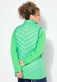 Chaleco acolchado de color verde claro con un patrón de espiga en la parte posterior, que presenta un alto cuello y mangas largas, combinado con una parte superior verde a juego.