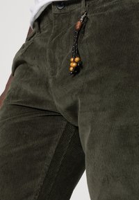 Olivgrön kordfloddsbyxor med en strukturerad ribbad yta. Har en dragkedjestängning och en dekorativ tassel av svarta och träperlor vid midjan.