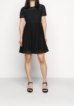 Femme portant une robe en dentelle noire longueur genoux avec des manches courtes et des talons noirs à bride cheville, debout devant un fond blanc uni.