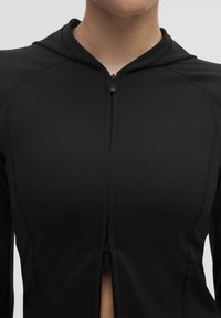 Jacheta sport neagră, cu fermoar și glugă, confecționată dintr-un material neted și elastic, cu cusături plate și buzunare laterale cu fermoar pentru funcționalitate.