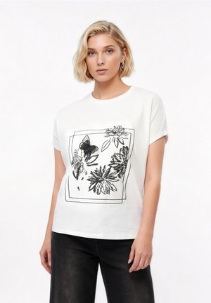 Νεαρή γυναίκα με κοντά ξανθά μαλλιά φοράει λευκό μπλουζάκι με μαύρο σχέδιο λουλουδιών και πεταλούδων, συνδυασμένο με μαύρο παντελόνι.