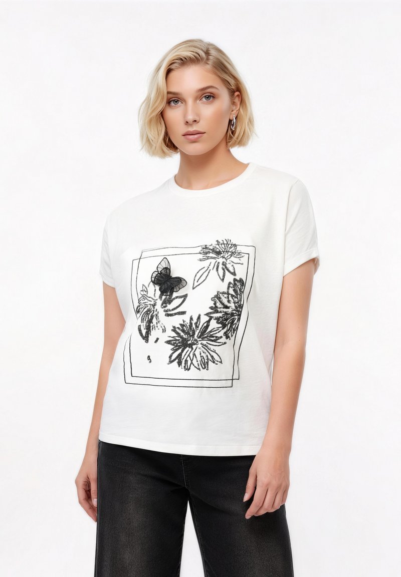 Νεαρή γυναίκα με κοντά ξανθά μαλλιά φοράει λευκό μπλουζάκι με μαύρο σχέδιο λουλουδιών και πεταλούδων, συνδυασμένο με μαύρο παντελόνι.