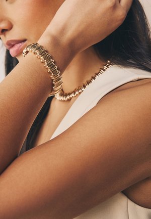 Woman wearing matching gold bracelet and necklace with horizontally stacked, rounded rectangular links, dressed in a white sleeveless top.