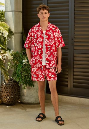 Mladý muž stojí oblečený v červenej kvetinovej havajskej košeli a zodpovedajúcich šortkách s potlačou Snoopyho, čierne sandále a biele tielko.