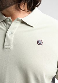 Ljusgrön polo med texturerad yta, med krage, två knappar och en rund, flerfärgad logotyp på bröstet.
