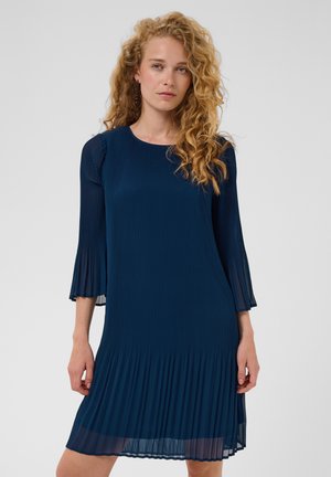 Femme aux cheveux blonds bouclés portant une robe plissée bleu marine à manches trois-quarts sur un fond gris uni.